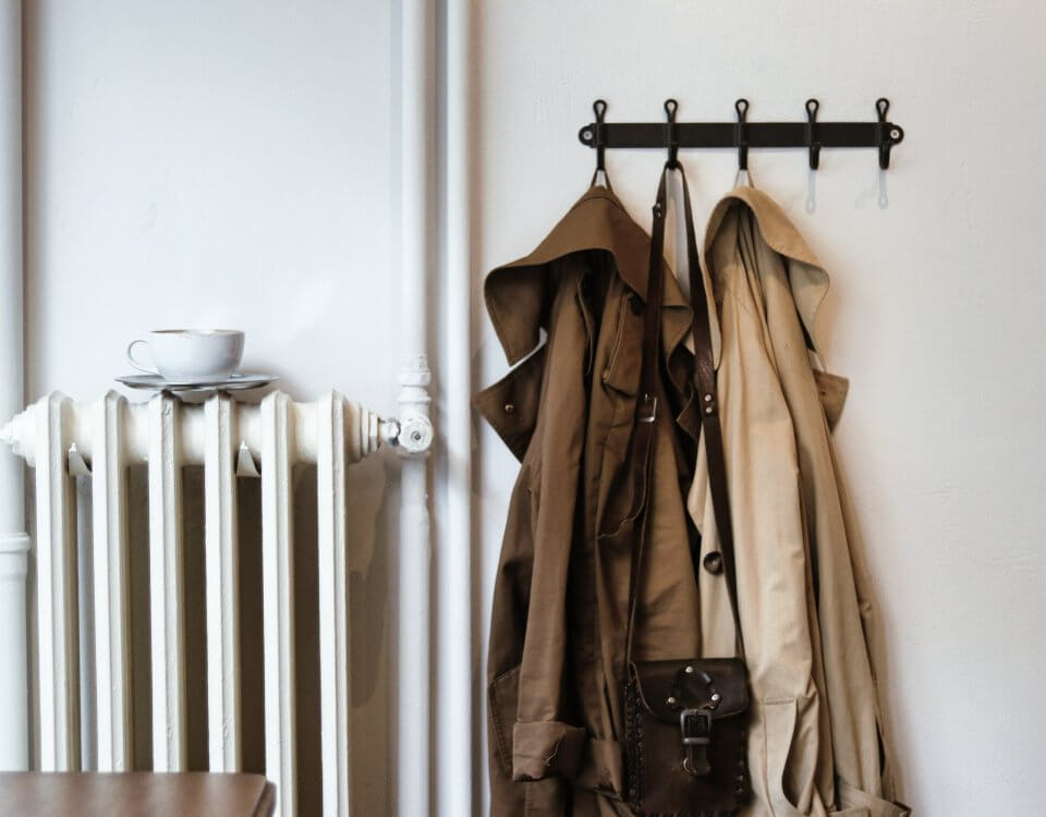 A radiator for heating on a wall next to a coat rack holding two coats and a handbag.